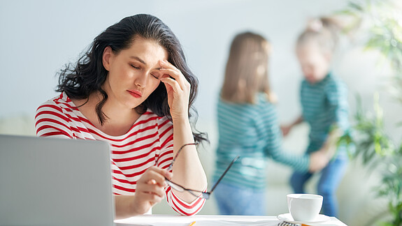 Sorgearbeit fair teilen Eine dunkelhaarige Frau arbeitet am Laptop und reibt sich gestresst die Augen, im Hintergrund spielen zwei kleine Mädchen