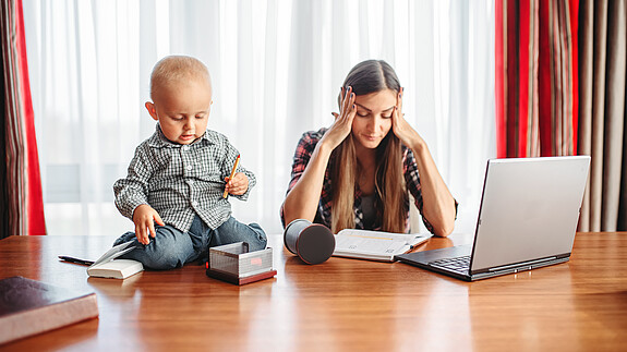 Erwerbsarbeit und private Care-Arbeit müssen zusammen gedacht werden Eine sichtlich gestresste Mutter versucht zu Hause am Laptop zu arbeiten, während neben ihr auf dem Schreibtisch ein Kleinkind sitzt und mit Büroutensilien spielt.