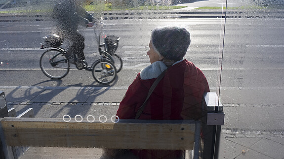 ÖPNV Eine ältere Dame in winterlicher Bekleidung sitzt an einer Bushaltestelle und wartet.