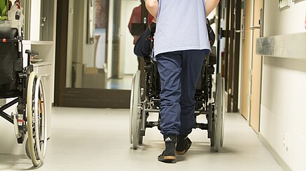 Eine Pflegekraft, von hinten zu sehen, schiebt einen älterne Menschen im Rollstuhl über einen Flur .