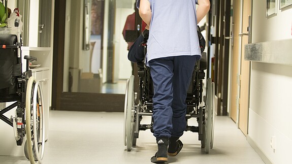 Pflege Eine Pflegekraft, von hinten zu sehen, schiebt einen älterne Menschen im Rollstuhl über einen Flur .
