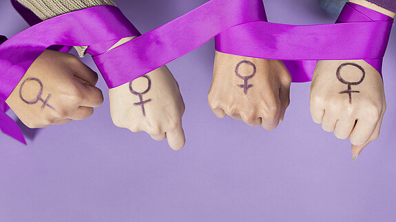 Vier Frauenhände mit einem aufgemalten Frauen-Symbol, ein violettes Band ist um die Handgelenke gelegt