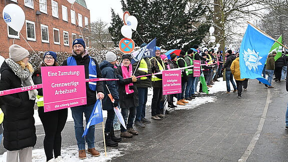 Menschen mit pinken Schildern und Warnwesten stehen als Menschenkette entlang einer langen Straße