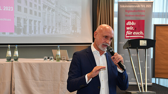 Ein Mann steht vor einer Leinwand mit einem Mikrofon in der Hand 