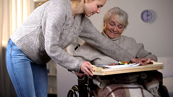 Entgeltersatzleistung für Pflegende - Silberbach: „Besser spät als nie!“ Das Foto zeigt eine Frau, die sich um eine Angehörige kümmert. Sie serviert Essen auf ein Tablett am Rollstuhl.