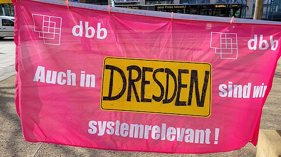 Ein Banner wird in die Kamera gehalten, auf dem steht "Auch in Dresden sind wir systemrelevant.""