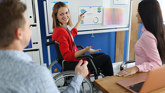 Inklusiver Arbeitsmarkt Ein im Rollstuhl sitzende Frau hält vor Kolleginnen und Kollegen einen Vortrag und zeigt auf ein Flipchart, auf dem diverse Grafiken angepinnt sind.