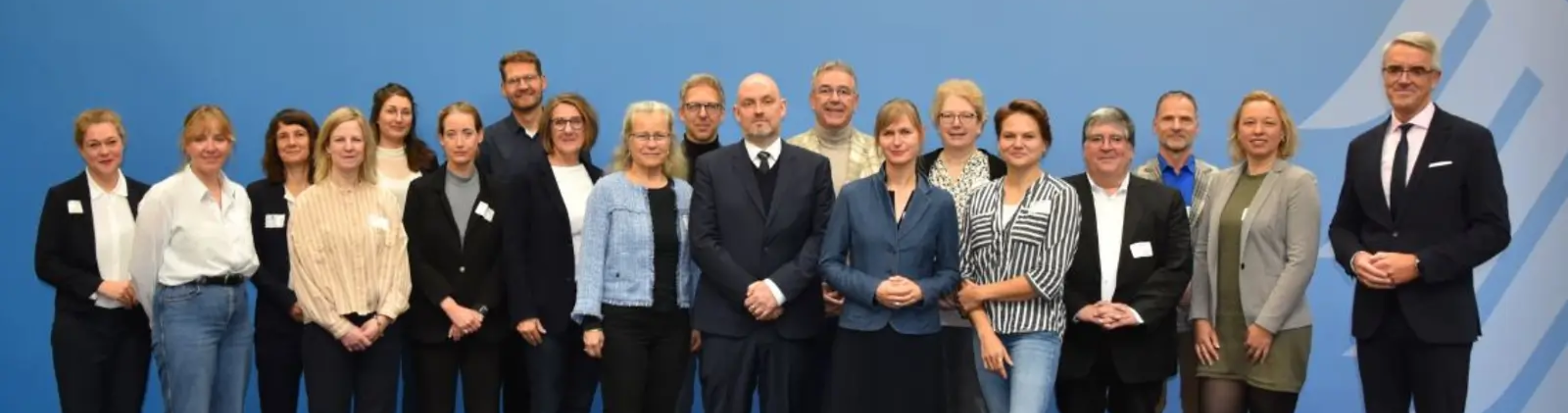 Eine große Gruppe Menschen steht vor einer hellblauen Wand und blickt in die Kamera.